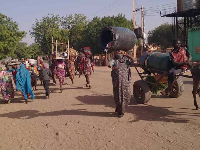 Families fleeing the violence in Western Darfur. A woman carries a barrel over her head next to a man with cart. Behind them are a multitude of families carrying their possessions.